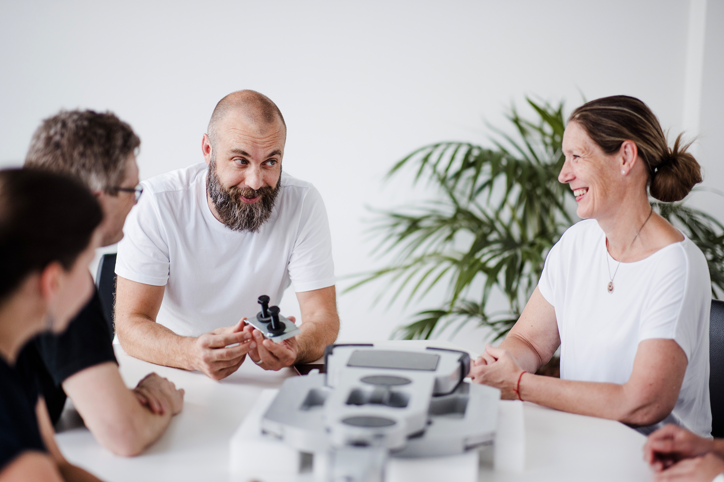 Gruppe von Personen diskutiert ein technisches Modell bei einem Meeting. Eine Gruppe von vier Personen sitzt um einen Tisch und bespricht gemeinsam ein technisches Modell. Ein Mann in einem weißen T-Shirt hält ein Bauteil in der Hand und erklärt es den anderen. Die Gruppe scheint in ein konstruktives und freundliches Gespräch vertieft zu sein. Im Hintergrund befindet sich leicht unscharf eine Pflanze.