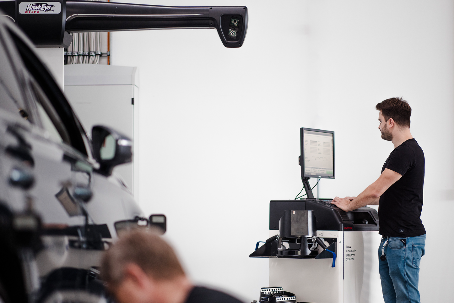 Mann arbeitet an einem Diagnose-System in einer Werkstatt. Ein Mann steht an einem Computerterminal in einer Werkstatt und arbeitet mit einem Diagnose-System, während ein Auto im Vordergrund teilweise sichtbar ist. Der Mann trägt ein schwarzes T-Shirt und konzentriert sich auf den Bildschirm.