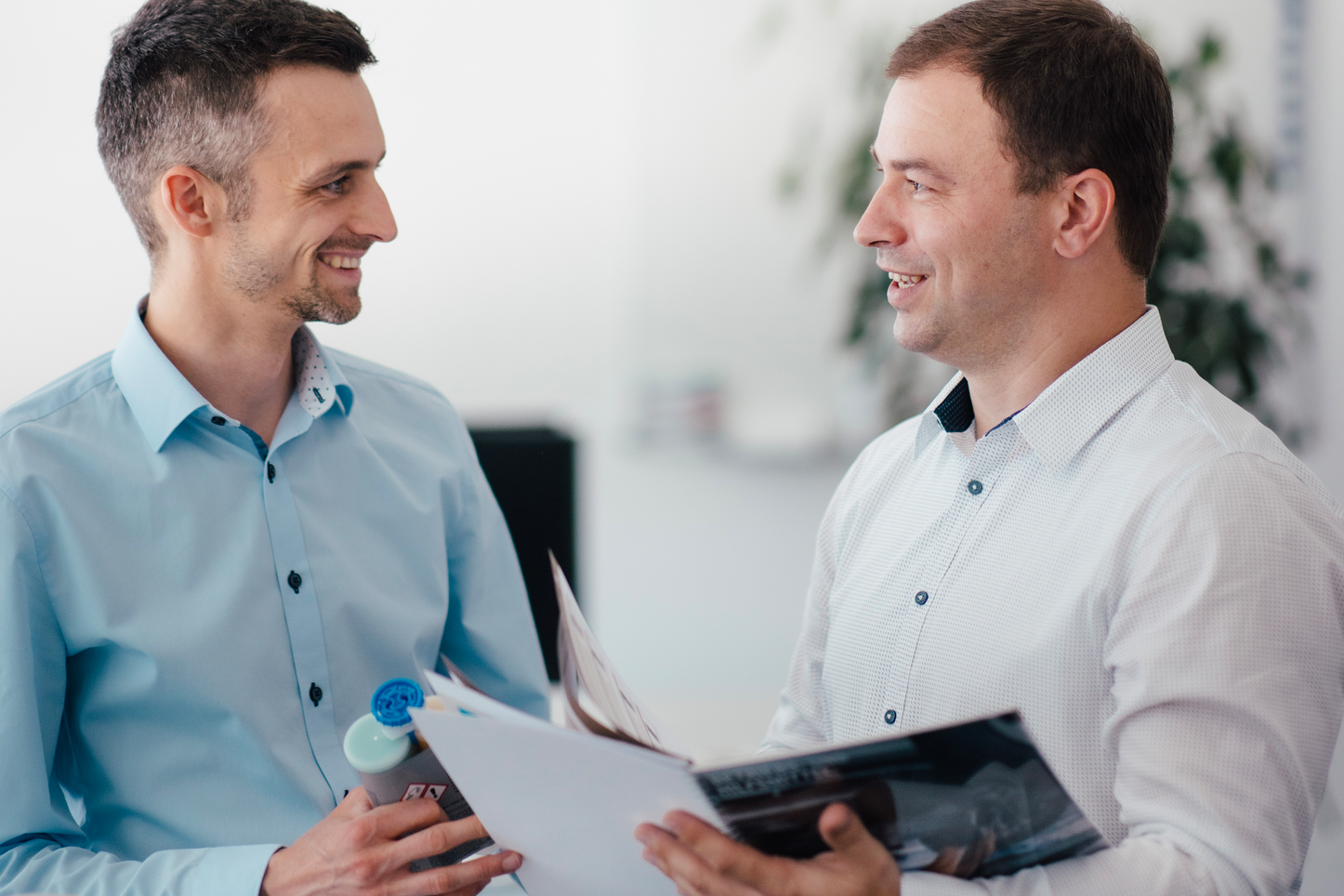 Austausch über Technische Dokumentation  Zwei Mitarbeiter unterhalten sich lächelnd in einem Büro und tauschen technische Dokumentationen aus. Einer der Mitarbeiter hält ein Produkt in der Hand, während der andere eine Mappe mit Dokumenten durchblättert.
