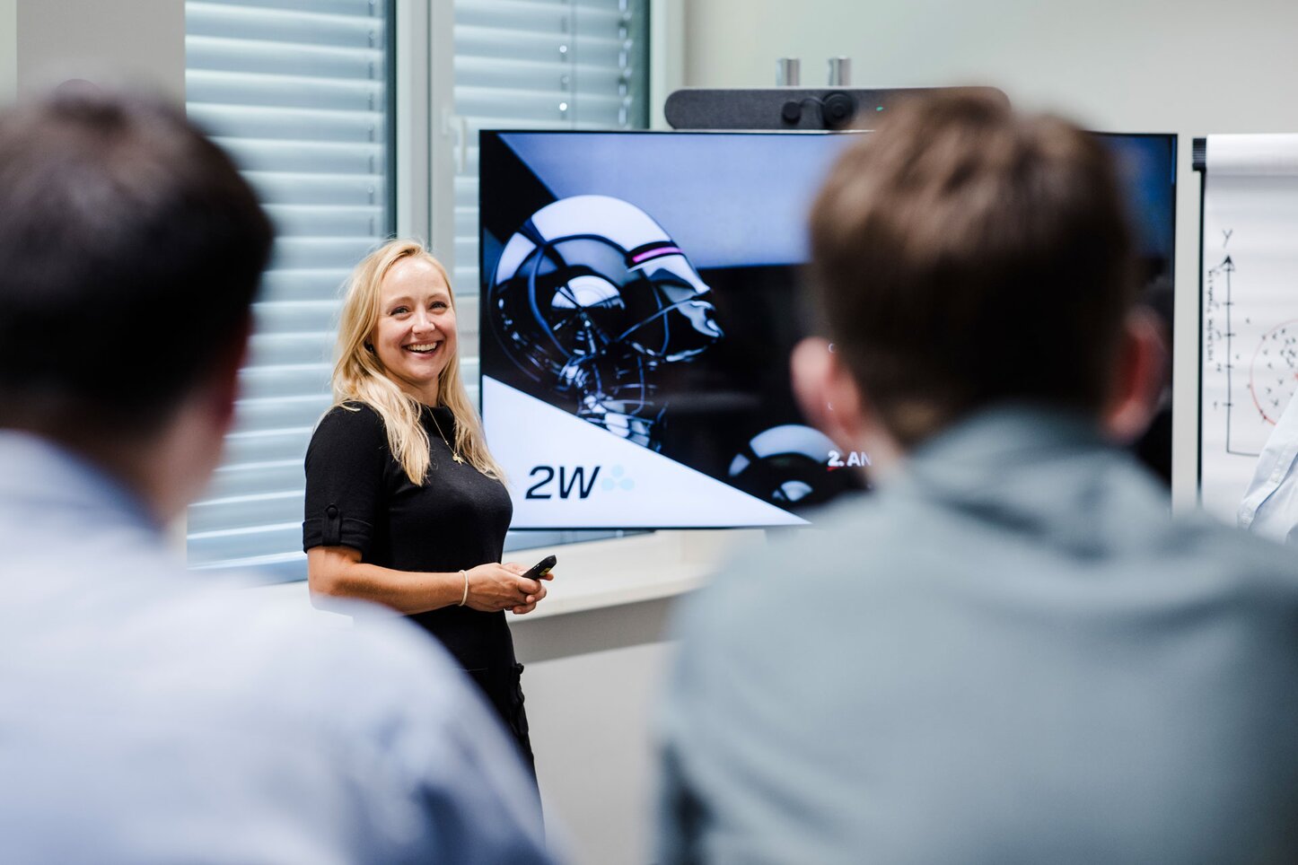 Frau hält eine Präsentation über technische Inhalte vor einem Bildschirm. Eine Frau hält eine Präsentation vor einem Bildschirm, der technische Grafiken zeigt, und lächelt dabei in Richtung des Publikums. Sie hält einen Präsentationsklicker in der Hand. Im Vordergrund sind unscharf zwei Zuhörer zu sehen, die der Präsentation folgen. Im Hintergrund ist eine Flipchart mit Diagrammen zu erkennen.
