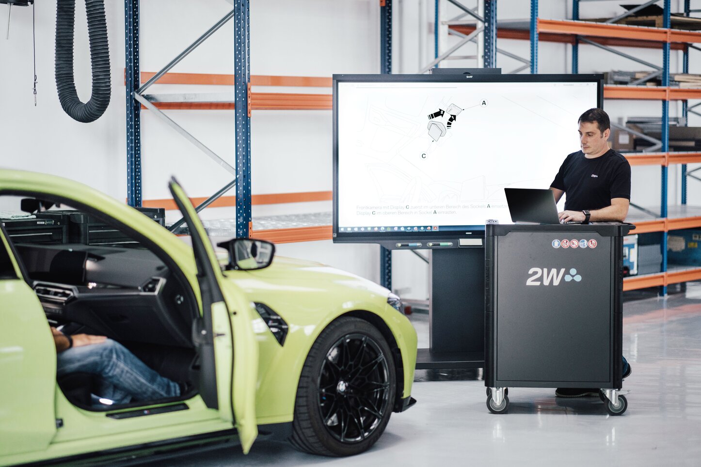 Datenanalyse und Prozessautomatisierung für die Homologation von Fahrzeugsystemen Ein Mann arbeitet an einem Laptop in einer Werkstattumgebung vor einem Bildschirm mit einer technischen Zeichnung. Ein grünes Auto mit offener Tür steht im Vordergrund. Der Fokus liegt auf Datenanalyse und Automatisierung von Homologationsprozessen.