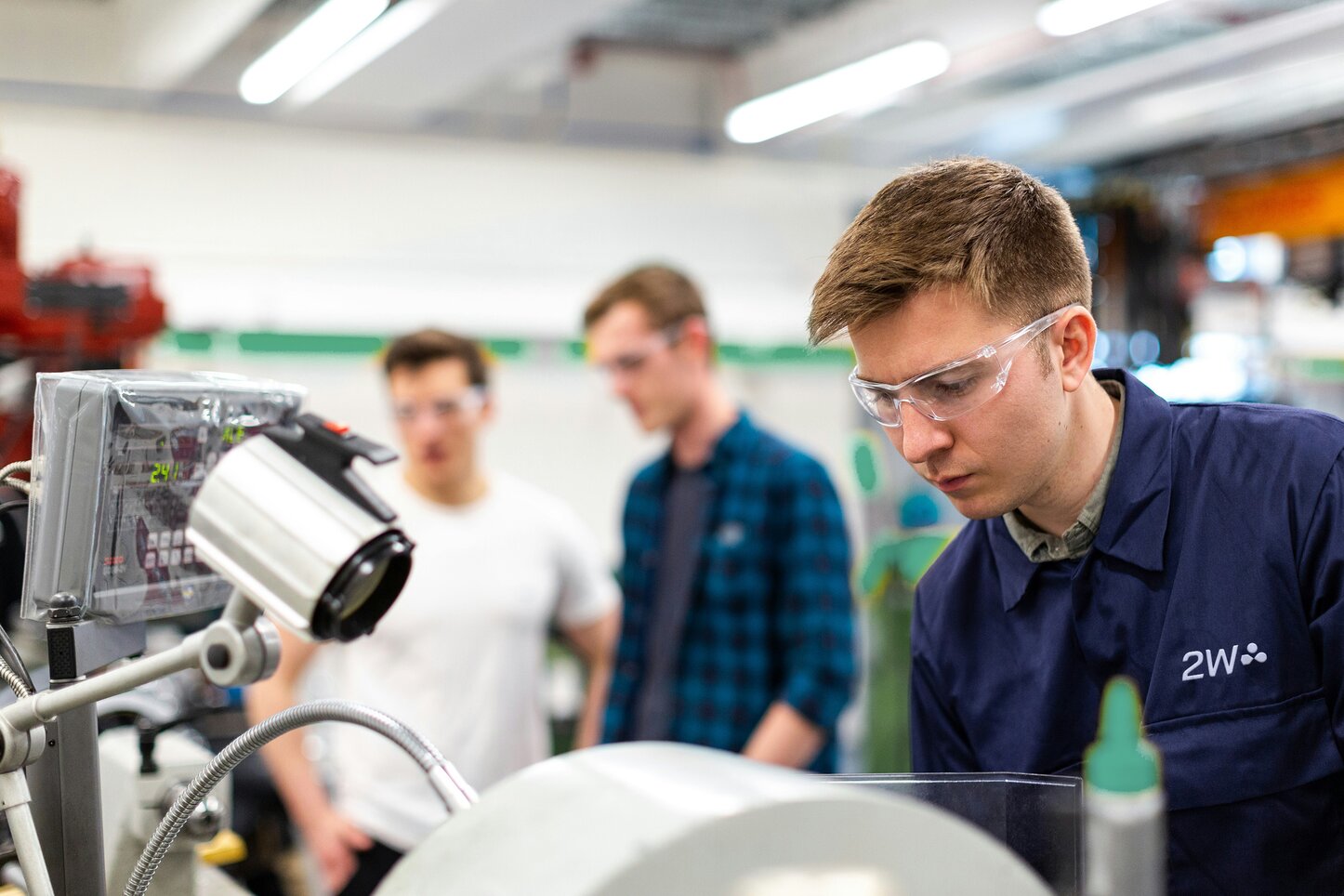 Junger Mann arbeitet an einer Maschine in einer Werkstatt, während zwei Kollegen im Hintergrund zuschauen. Ein junger Mann in Schutzbrille und 2W-Arbeitskleidung arbeitet konzentriert an einer Maschine in einer Werkstatt. Im Vordergrund ist die Maschine mit einem Monitor und technischen Geräten zu sehen. Im Hintergrund stehen zwei weitere Personen, die ebenfalls Schutzbrillen tragen und die Szene beobachten oder an einem anderen Projekt arbeiten.
