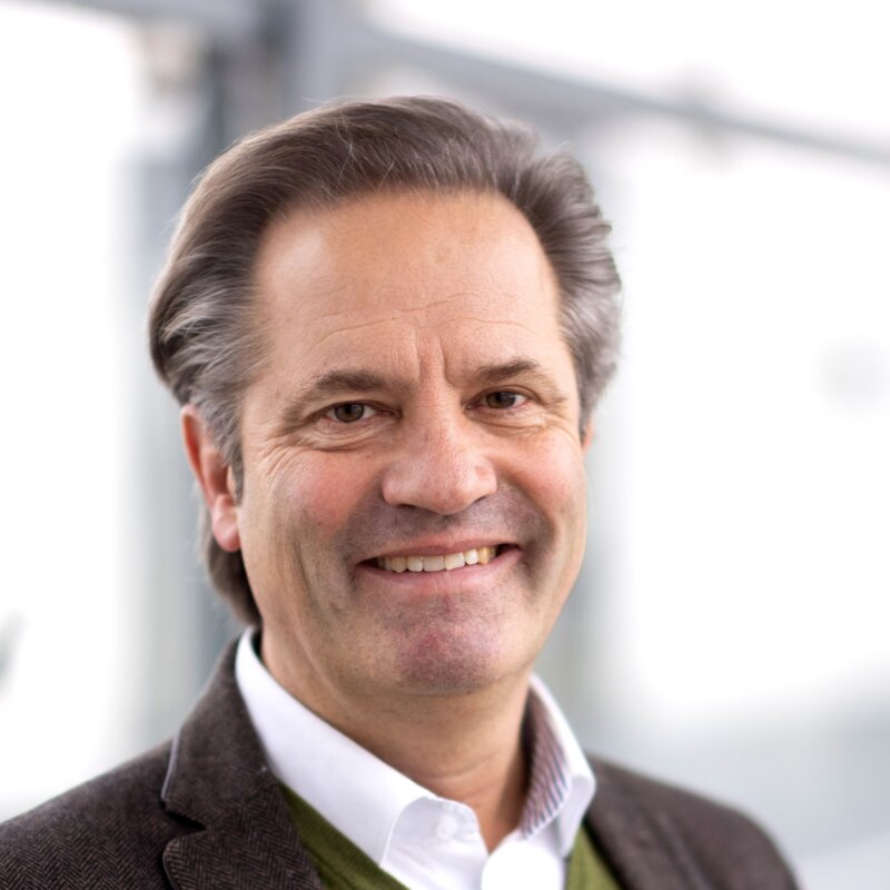 Portraitfoto des Geschäftsführers Michael Süssl Ein Mann mit mittellangem, leicht ergrautem Haar sitzt in einem hellen Büro und blickt freundlich in die Kamera. Er trägt ein weißes Hemd, das eine professionelle Ausstrahlung vermittelt. Der Hintergrund ist unscharf und zeigt große Fenster, durch die Tageslicht einfällt.