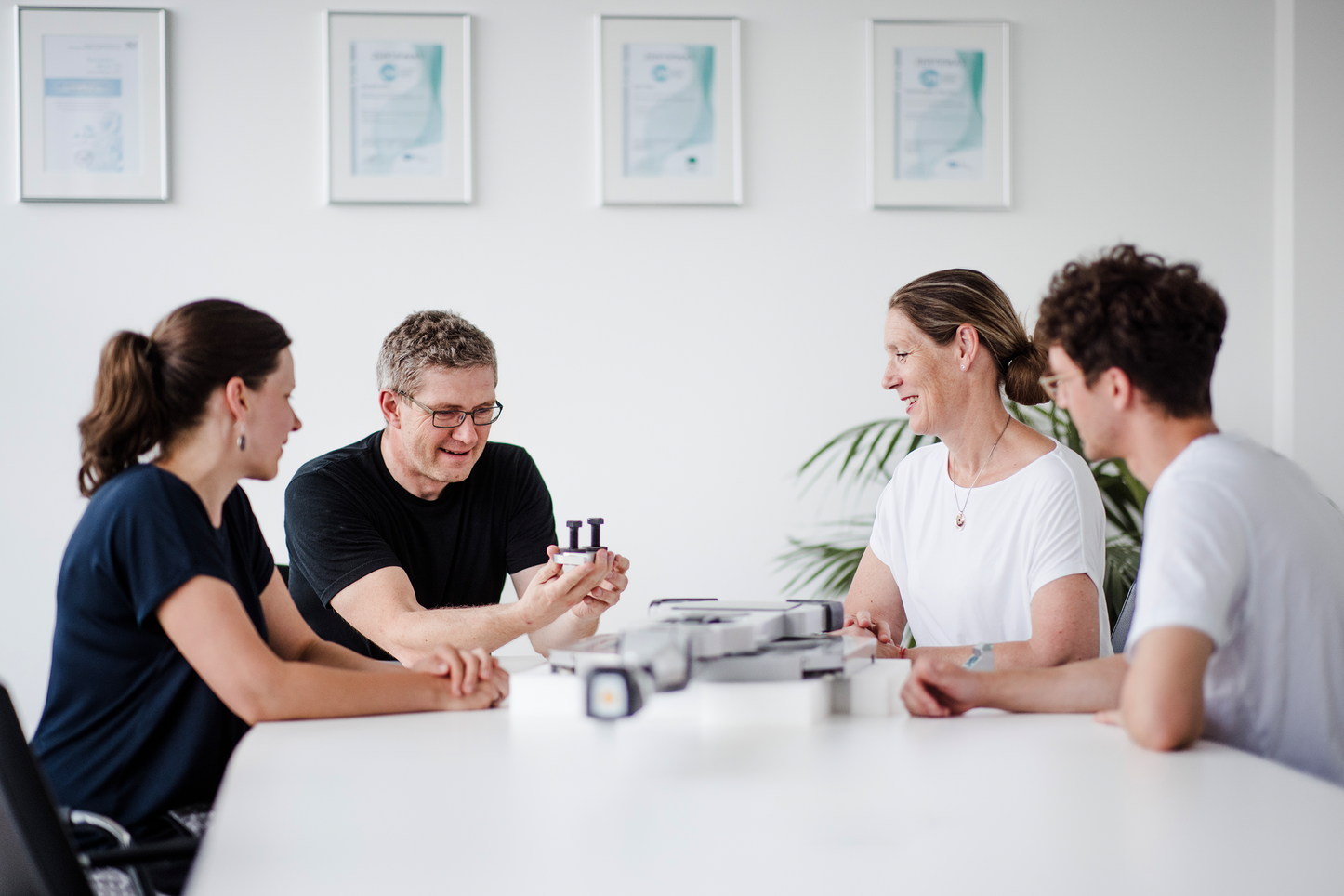 Team-Meeting mit Produktbesprechung Vier Personen sitzen in einem Besprechungsraum an einem Tisch und unterhalten sich lächelnd. Eine Person hält ein kleines Bauteil in den Händen und zeigt es den anderen, die interessiert zuschauen. Auf dem Tisch liegen weitere technische Geräte. Im Hintergrund hängen Zertifikate an der Wand.