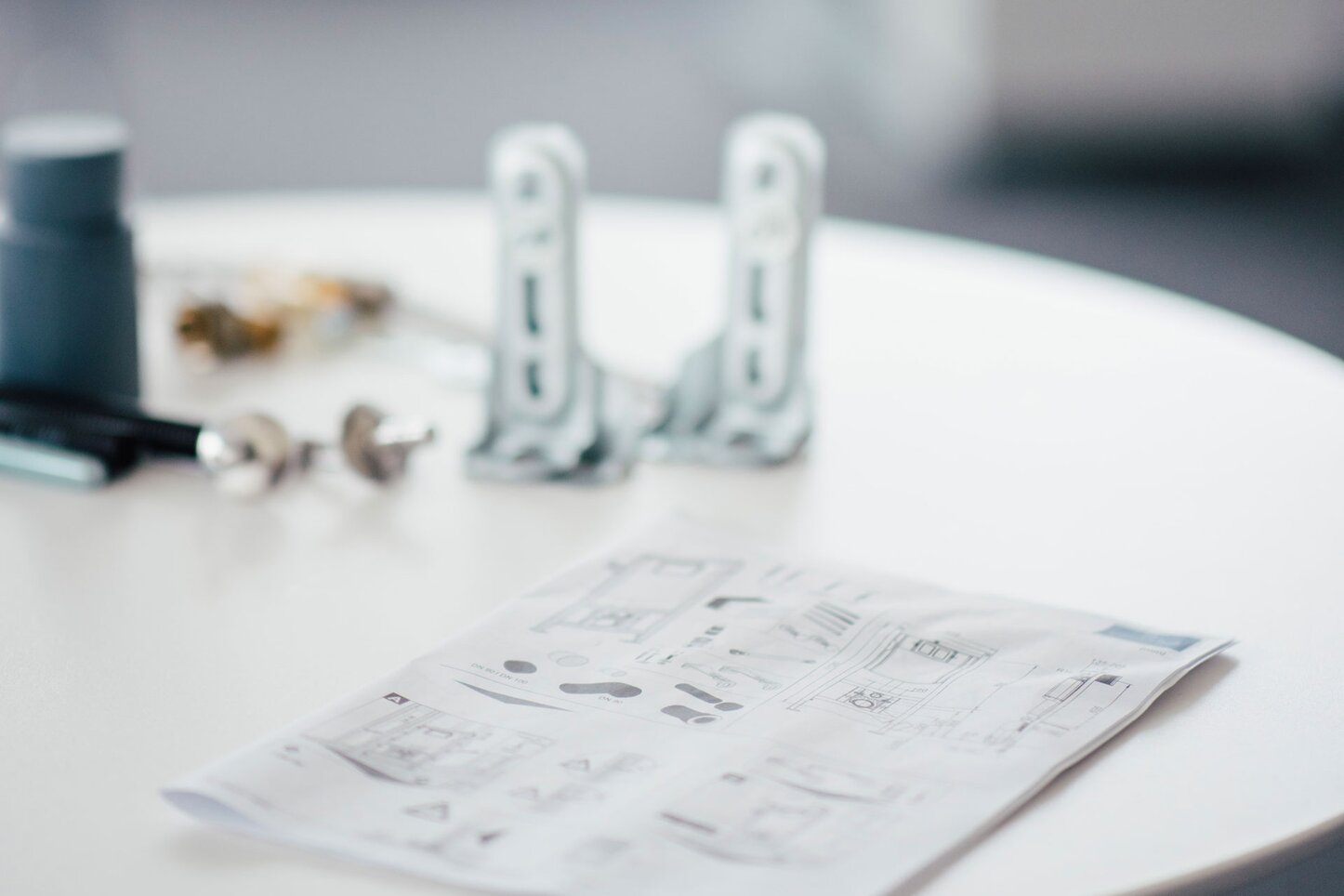 Tisch mit Metallbauteilen und Montageanleitung in einem Büro. Ein runder Tisch in einem Büro mit mehreren Metallbauteilen und einer ausgebreiteten Montageanleitung im Vordergrund. Im Hintergrund sind unscharf eine Pflanze und weitere Büroelemente zu erkennen.