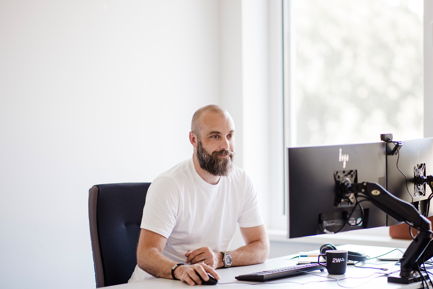 Mitarbeiter bei der Arbeit am Schreibtisch mit Doppelbildschirm Ein Mann mit Bart sitzt an einem Schreibtisch vor zwei Monitoren und bedient die Computermaus. Er trägt ein weißes T-Shirt und wirkt konzentriert.