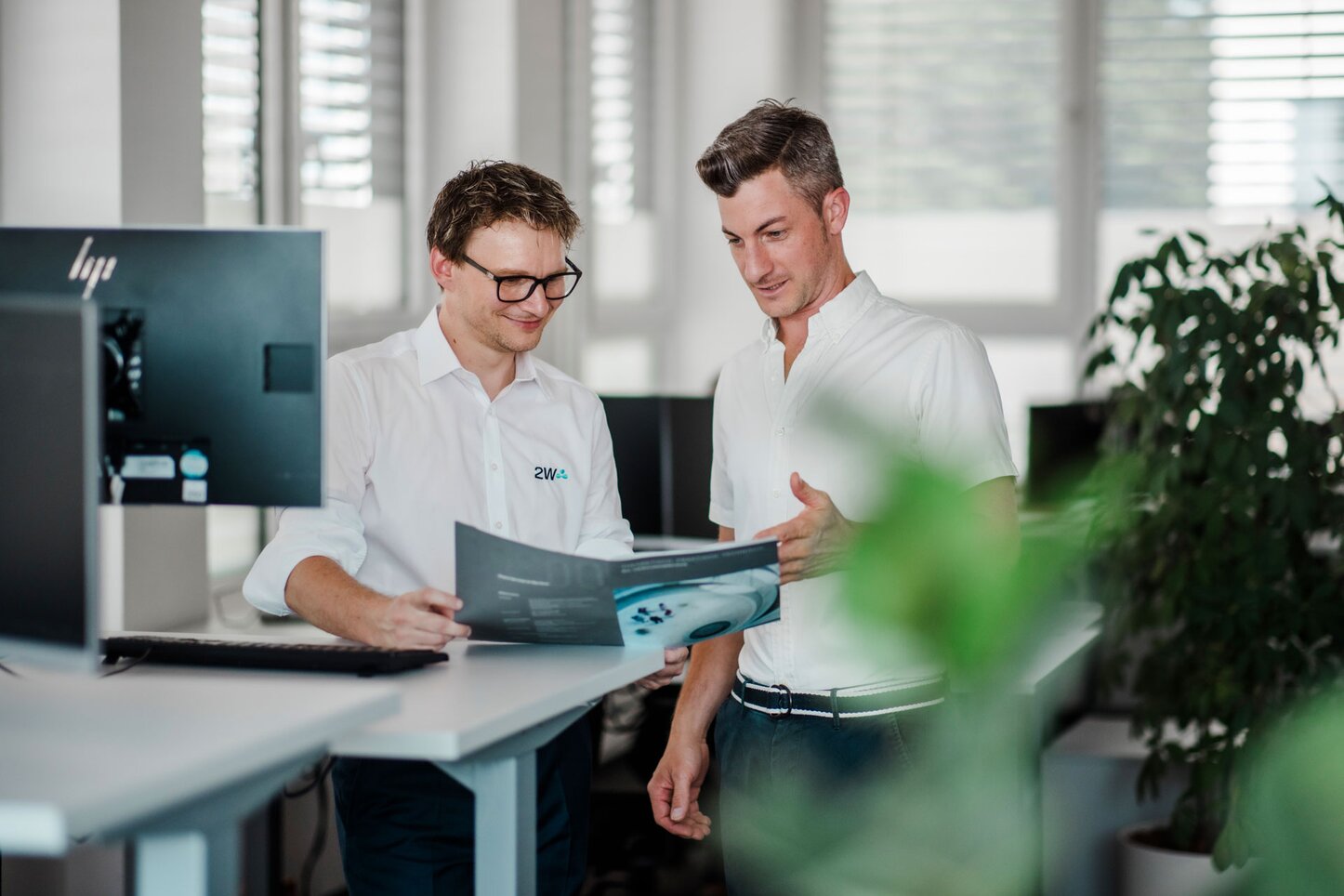 Zwei Männer besprechen eine technische Broschüre in einem modernen Büro. Zwei Männer stehen in einem modernen Büro vor einem Computerbildschirm und betrachten gemeinsam eine technische Broschüre. Beide lächeln und diskutieren offenbar über den Inhalt des Dokuments. Im Vordergrund sind einige Pflanzen zu sehen.