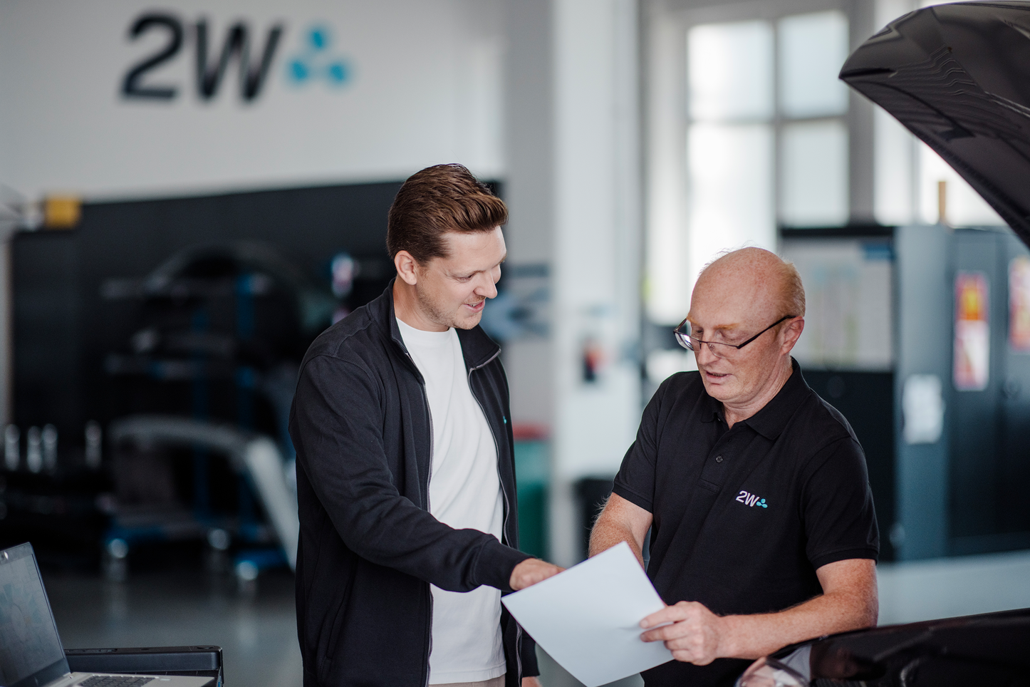 Zwei Männer besprechen ein Dokument in einer Werkstatt. Zwei Männer stehen in einer Werkstatt und besprechen gemeinsam ein Dokument. Der Mann auf der linken Seite zeigt auf das Papier, während der ältere Mann auf der rechten Seite das Dokument in der Hand hält. Im Hintergrund sind Werkzeuge und Werkstatt-Ausrüstung zu sehen, und die Motorhaube eines Fahrzeugs ist geöffnet.