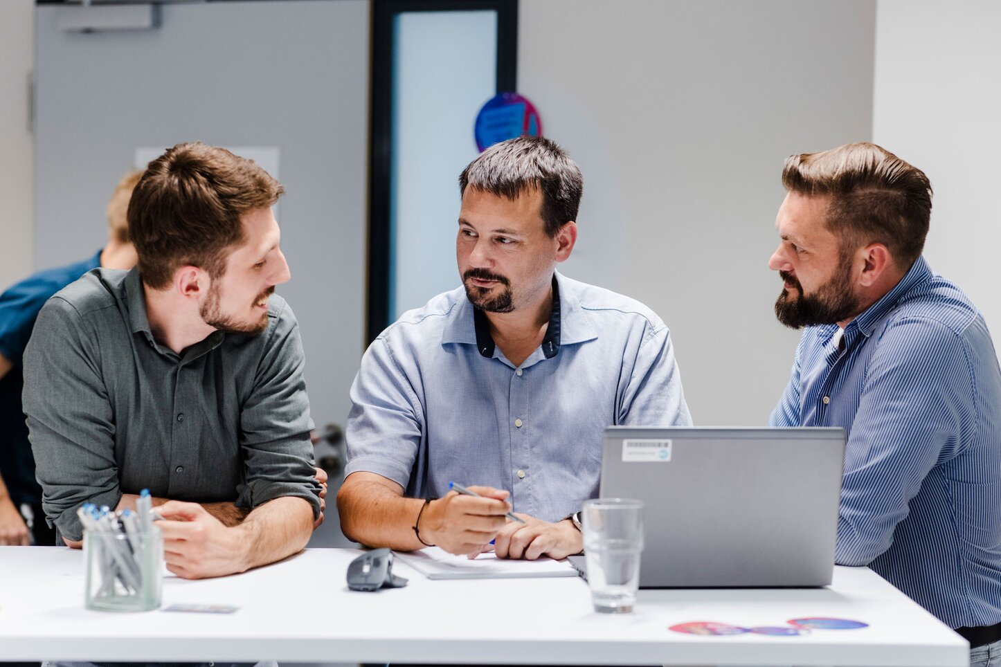 Geschäftstreffen: Drei Männer im Gespräch in einem modernen Büro Drei Männer in Geschäftskleidung sitzen um einen Tisch und diskutieren konzentriert miteinander. Auf dem Tisch befinden sich ein Laptop, ein Glas Wasser und ein Stiftbehälter. Einer der Männer hält einen Stift in der Hand und ein Notizblock liegt vor ihm. Alle drei sind in ein Gespräch vertieft, das eine professionelle und kooperative Atmosphäre vermittelt.