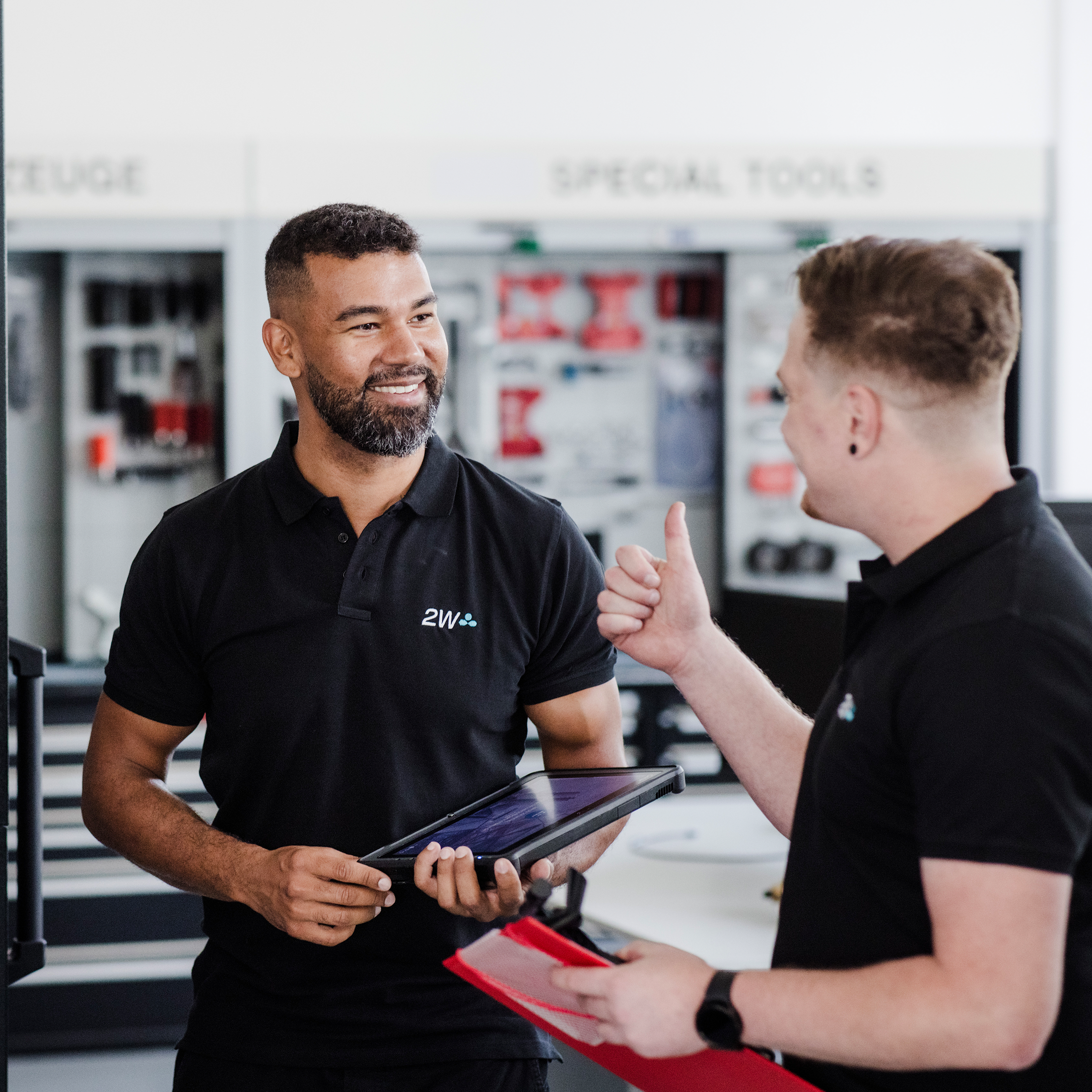 2W Mitarbeiter im Gespräch in der Werkstatt Zwei Techniker unterhalten sich in der Werkstatt. Einer hält ein Tablet, der andere eine Mappe. Im Hintergrund sind Werkzeuge und Ausrüstungen an der Wand zu sehen.