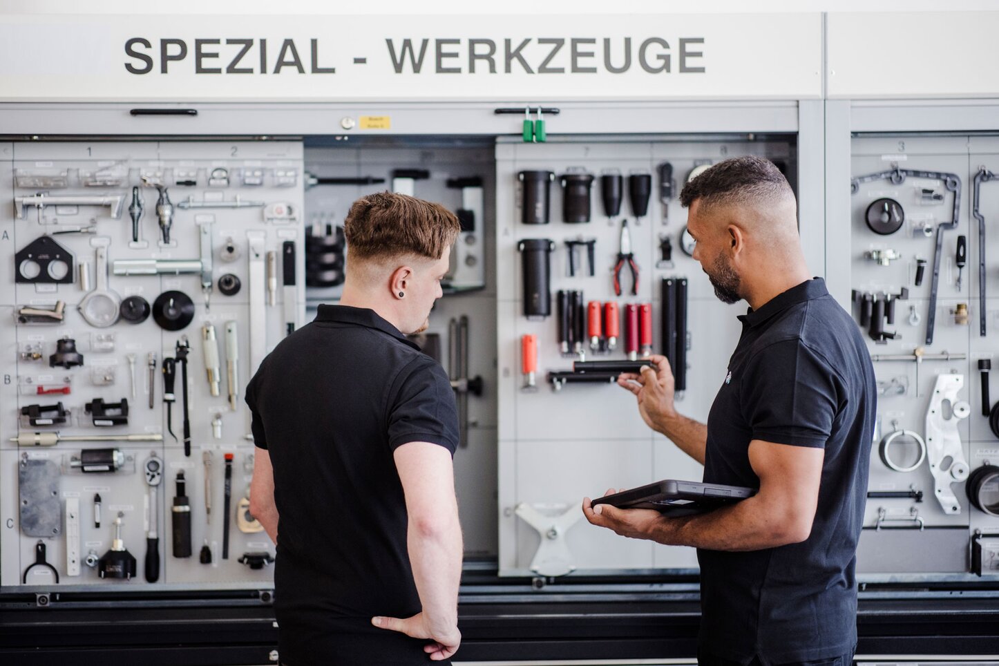 Zwei Männer begutachten Werkzeuge vor einem Schrank mit Spezial-Werkzeugen. Zwei Männer stehen vor einem Werkzeugschrank mit der Aufschrift "Spezial-Werkzeuge" und begutachten verschiedene Werkzeuge. Einer der Männer hält ein Tablet in der Hand, während der andere auf ein Werkzeug zeigt. Der Schrank ist mit einer Vielzahl von spezialisierten Werkzeugen bestückt, die ordentlich angeordnet sind.